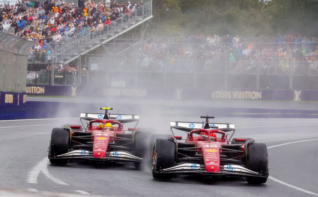 Ferrari (red car) number 16 Charles Leclerc and Ferrari (red car) number 44 Lewis Hamilton race side by side through a wet corner in front of a packed grandstand.