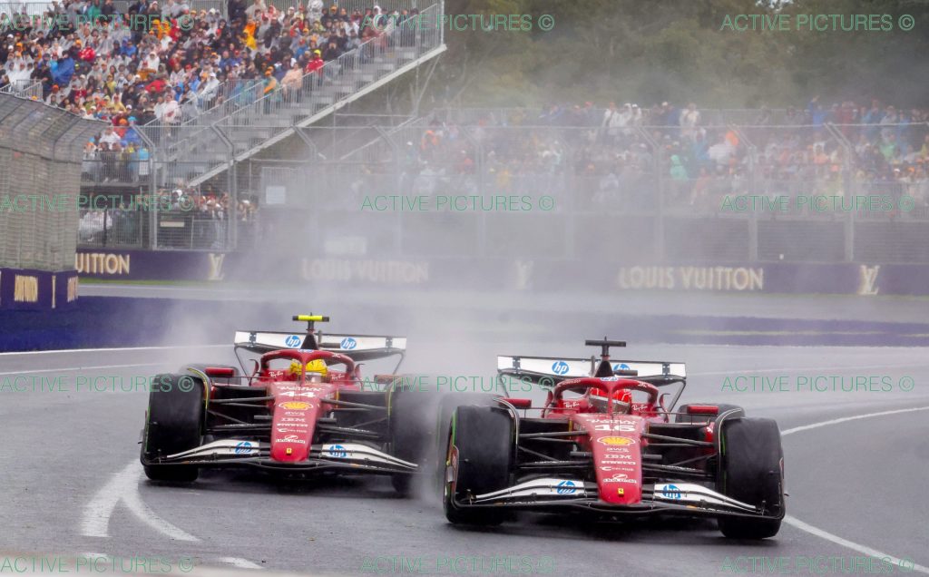 Ferrari (red car) number 16 Charles Leclerc and Ferrari (red car) number 44 Lewis Hamilton race side by side through a wet corner, kicking up spray in front of a packed grandstand.