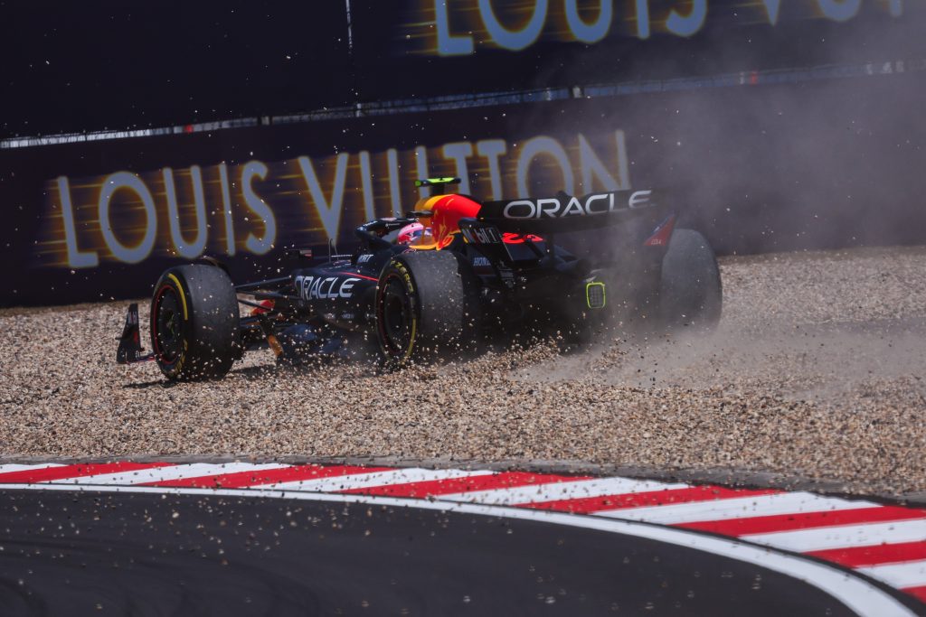 Red Bull (dark blue car) runs through the gravel trap, kicking up stones and dust during a dramatic moment at the circuit.