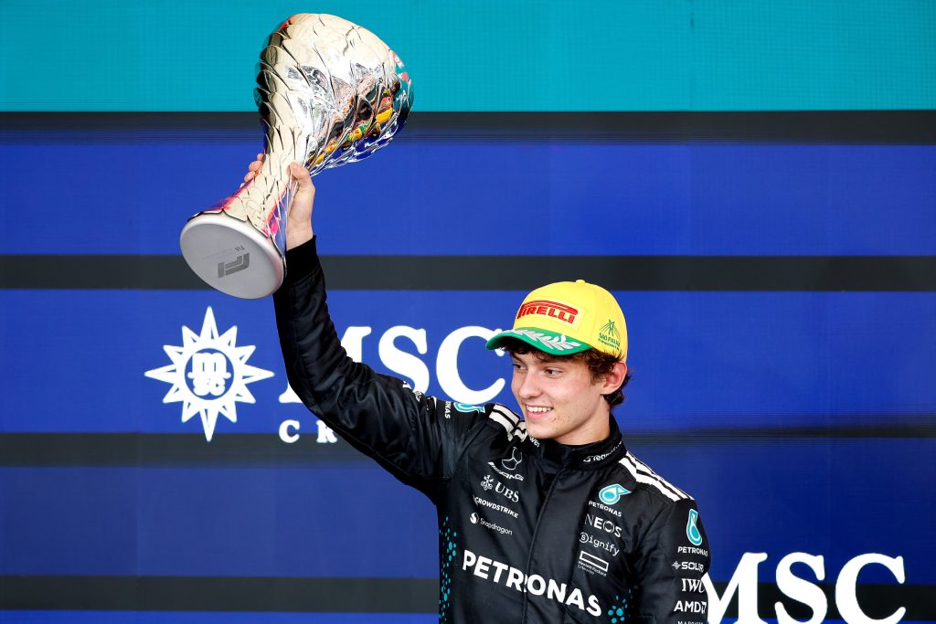Andrea Kimi Antonelli, wearing his Mercedes team race suit, celebrates on the podium holding a winner’s trophy aloft.