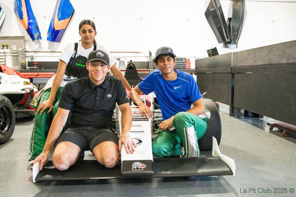 Three people pose around the nose and front wing of an open-wheel race car inside a workshop or garage, with other car parts and equipment visible in the background.
