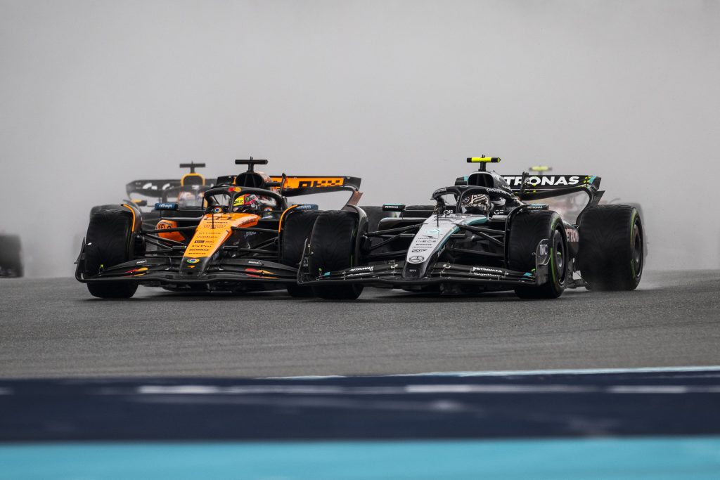Oscar Piastri’s McLaren #81 (orange car) runs side by side with Kimi Antonelli’s Mercedes #12 (silver and black car) on a damp straight, with the McLaren edging ahead slightly, while another McLaren (orange car) follows behind through the spray.