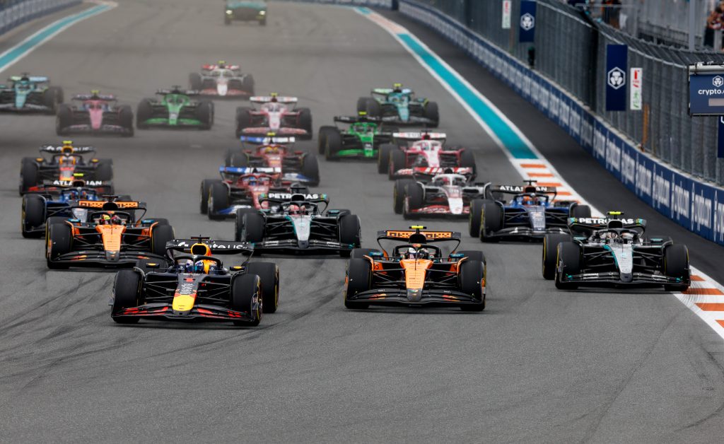 Max Verstappen’s Red Bull (dark blue car) #1 surges off the start between Lando Norris’s McLaren (orange car) #4 on the left and Oscar Piastri’s McLaren (orange car) #81 on the right, while George Russell’s Mercedes (silver and black car) #63 a...