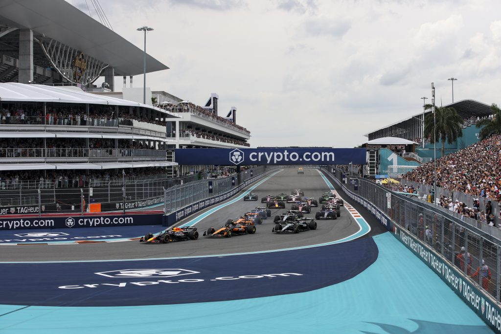 Max Verstappen’s Red Bull #1 (dark blue car with red and yellow details) leads into the opening left-hander, with Lando Norris’s McLaren #4 (orange car with black accents) challenging alongside, Andrea Kimi Antonelli’s Mercedes #12 (silver car wi...