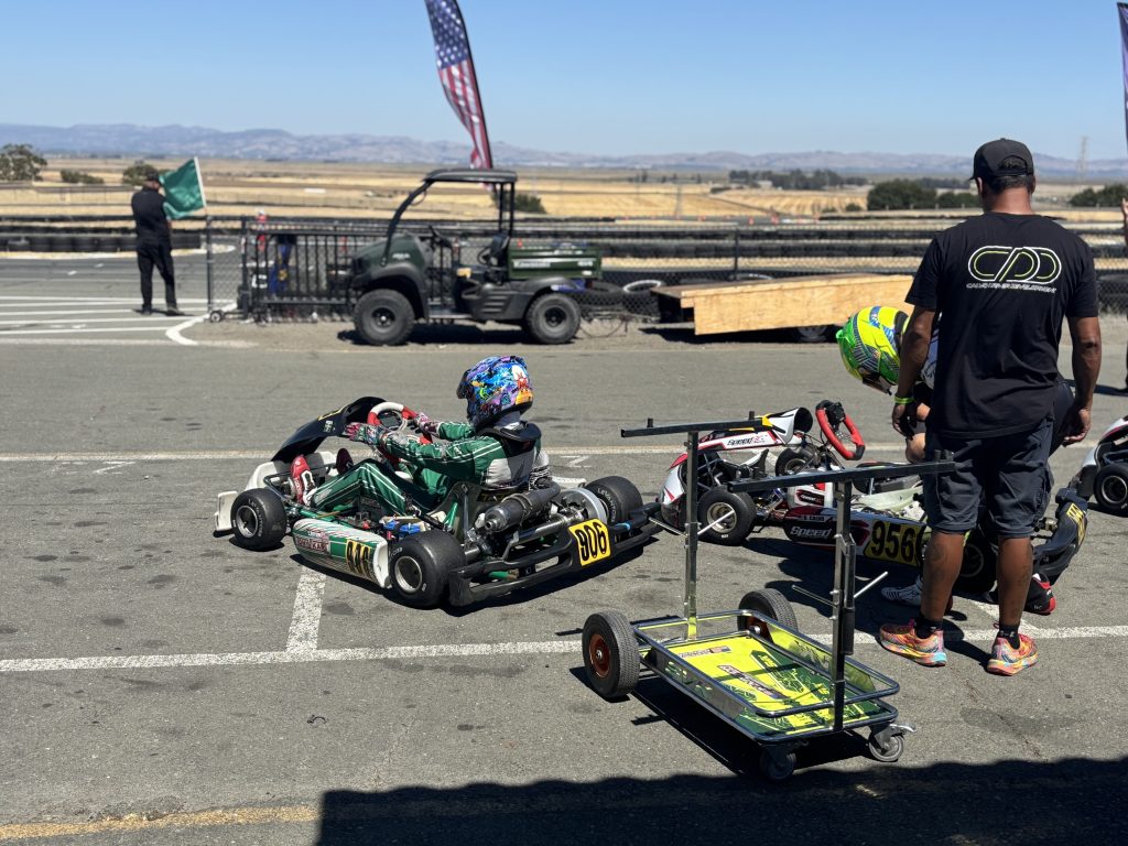 Two people in racing gear are beside small go-karts in a paddock area at a kart track, while a marshal in the background waves a green flag near a fence, a utility cart, and an American flag under a clear sky.