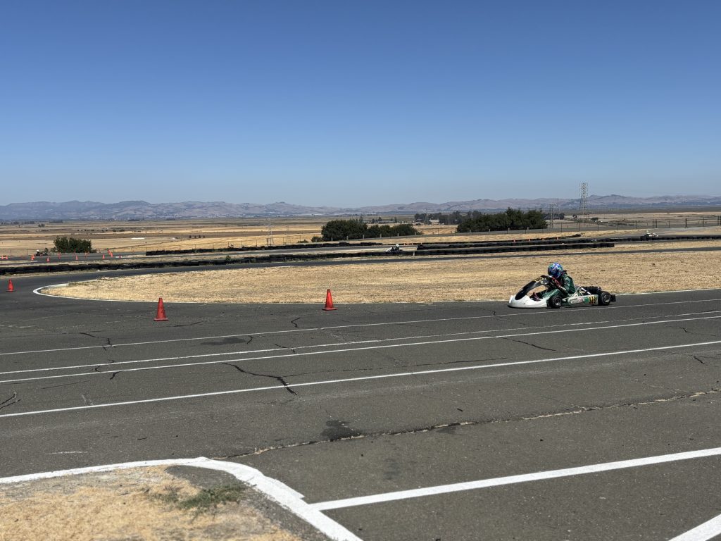 A go-kart driver rounds a paved track marked with orange cones, with a few other karts visible farther back against a dry, open landscape and distant hills under a clear blue sky.