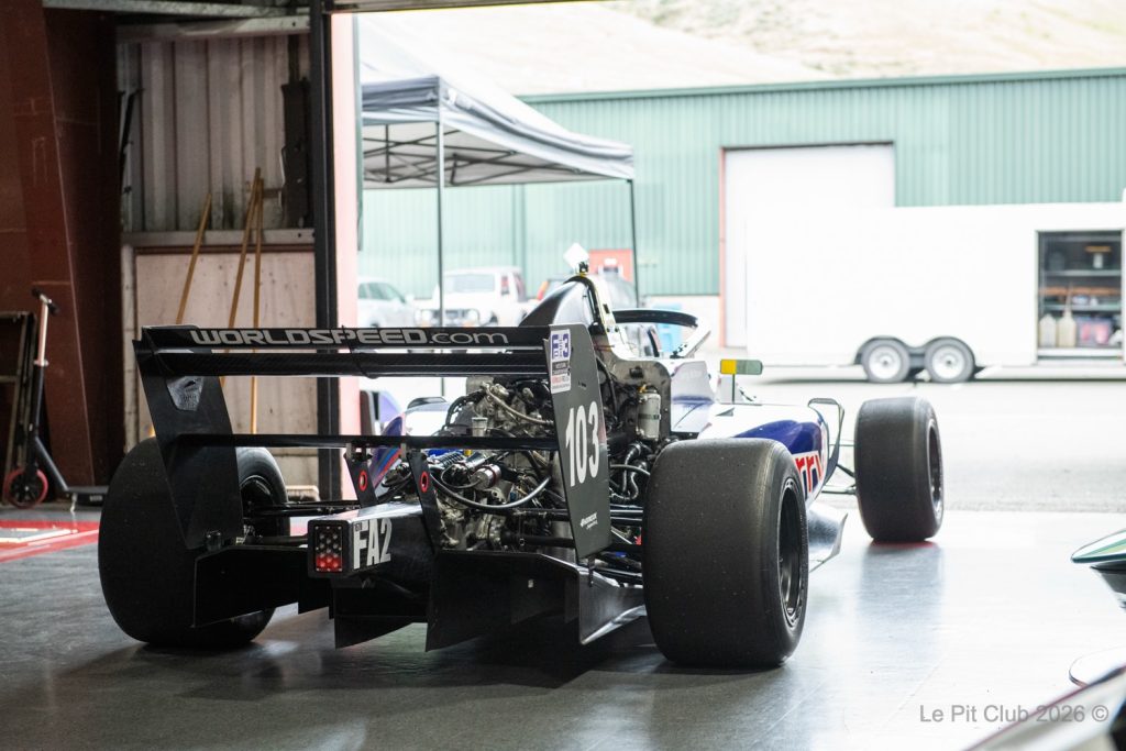 The rear of an open-wheel race car with a large rear wing and exposed engine is parked inside a garage, facing an open paddock area with a trailer and canopy outside.