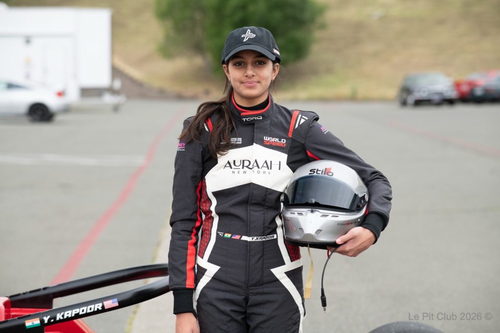 A person stands on a racetrack in a black, white, and red racing suit, holding a silver racing helmet beside an open-wheel race car. The suit shows “WORLD SPEED” and “AURAAI NEW YORK” branding, and the name “Y. KAPOOR” appears on both the s...