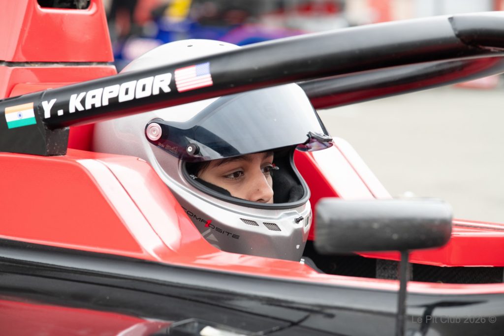 A close-up shows a helmeted driver seated in a red open-wheel race car, with the name “Y. Kapoor” and Indian and American flags printed on the halo above.