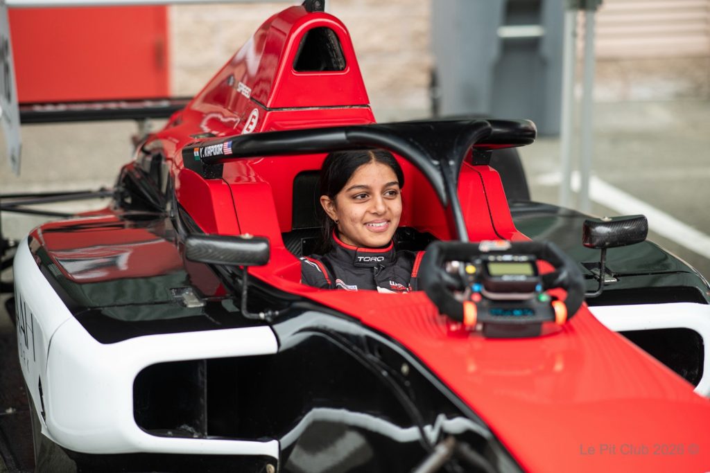 A smiling woman sits in the cockpit of a red, black, and white open-wheel race car, with the steering wheel mounted in front of her and a building interior blurred in the background.