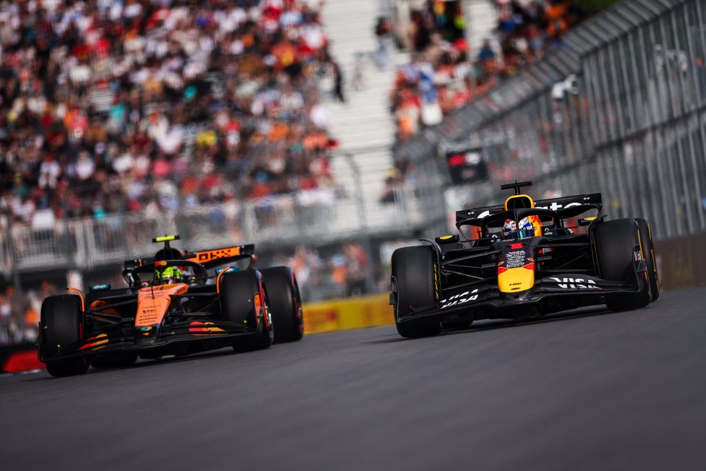 Max Verstappen’s Red Bull (dark blue with red and yellow details) #1 leads on the right side of the track, with Lando Norris’s McLaren (orange with black accents) #4 chasing from behind on the left as they run down the straight past packed grandsta...