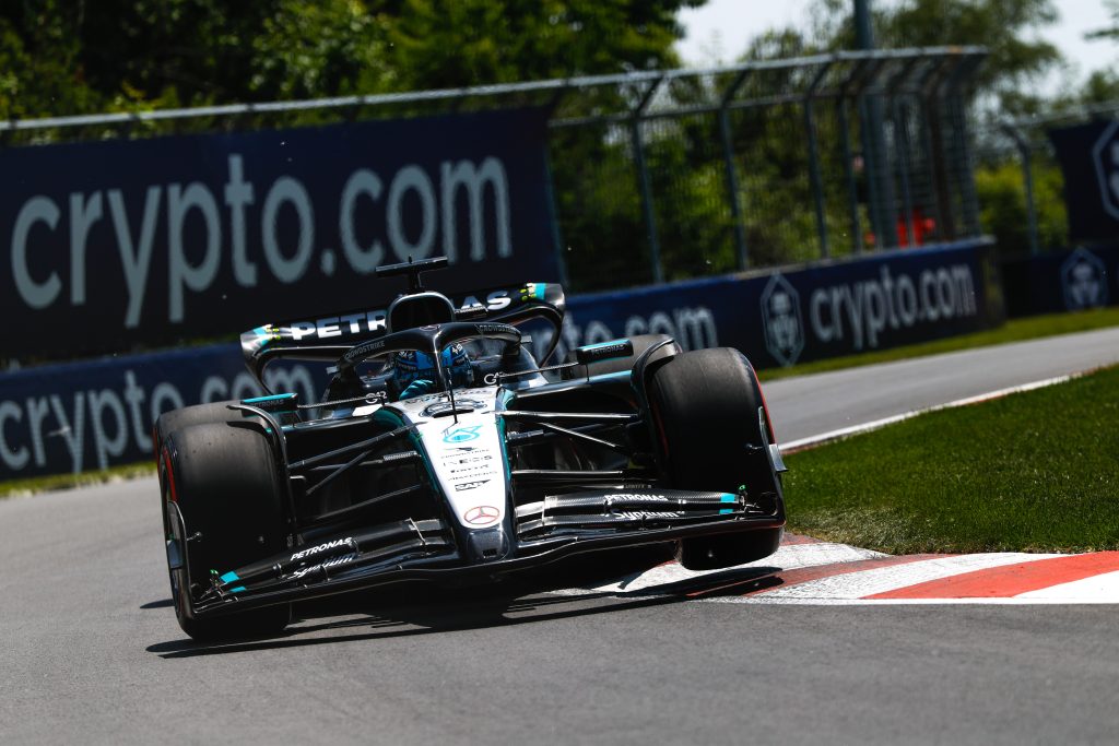 A Mercedes (silver with black elements and turquoise accents), George Russell’s #63, rides the red-and-white kerb aggressively through the corner with its inside wheels lifted slightly off the track.