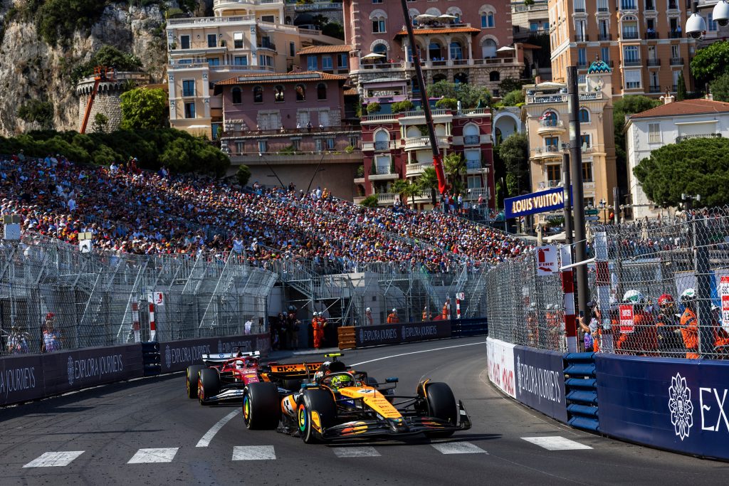 A McLaren (orange car), with Lando Norris visible in the cockpit, leads through the tight Monaco street section, while a Ferrari (red car) chases a few lengths behind.