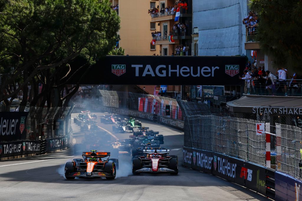 Oscar Piastri’s McLaren (orange car) #81 leads under braking with tire smoke billowing off the front-left, while a Ferrari (red car) runs alongside on the right and a Mercedes (silver and black car), an Aston Martin (green car), and an Alpine (blue a...