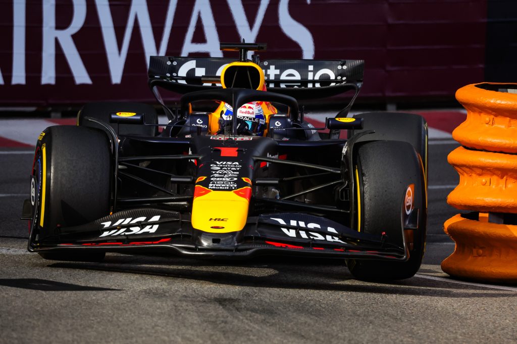 Max Verstappen’s Red Bull (dark blue car with red and yellow details) #1 charges through the Monaco street circuit alone, clipping the narrow line beside the orange barrier.