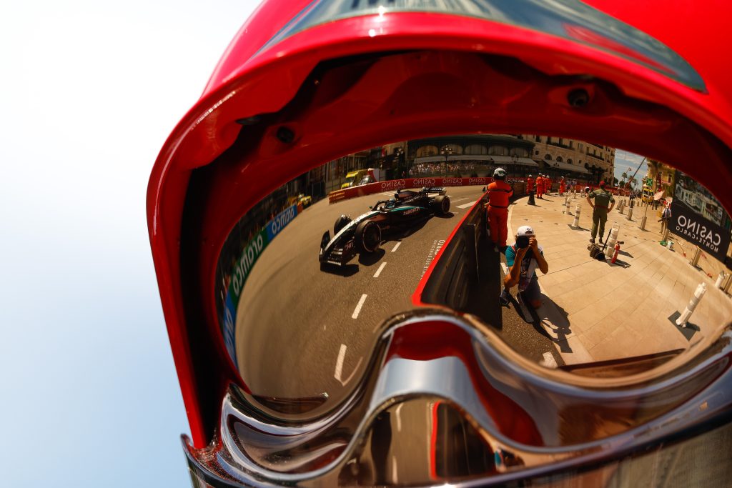 George Russell’s Mercedes (silver with black elements and turquoise accents car) appears in the mirrored visor as the #63 car sweeps through the Monaco street section, with another Mercedes (silver with black elements and turquoise accents car) follo...