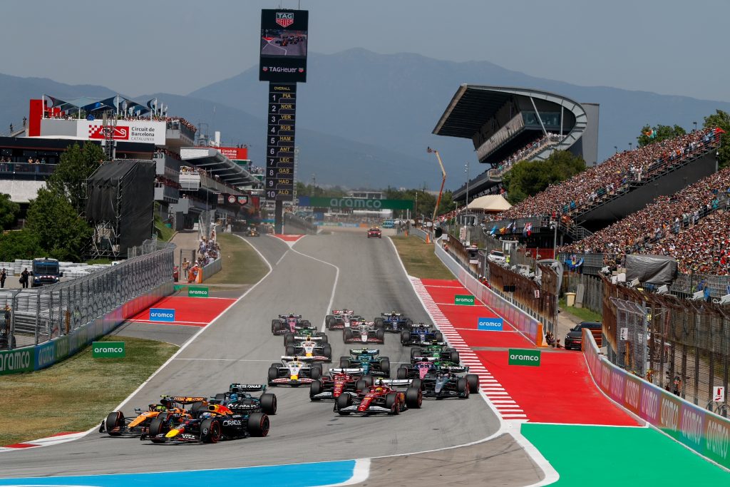 Max Verstappen’s Red Bull (dark blue car) #3 and Lando Norris’s McLaren (orange car) #1 charge side by side at the front into the opening corner, while Charles Leclerc’s Ferrari (red car) #16 and Lewis Hamilton’s Ferrari (red car) #44 follow cl...