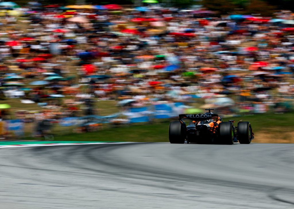A McLaren (orange car), driven by Oscar Piastri #81, runs alone over the crest of the circuit with the blurred hillside crowd in the background.