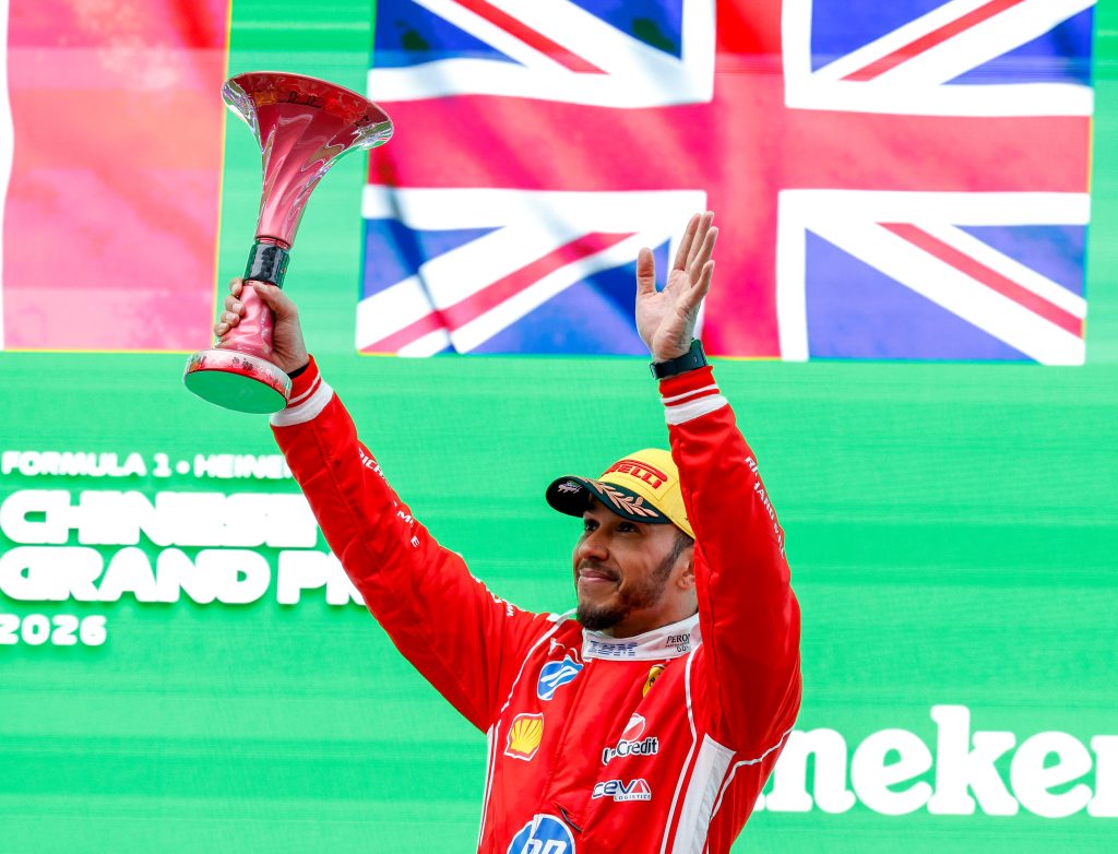 Ferrari driver Lewis Hamilton raises a trophy on the podium in front of a Union Jack backdrop.