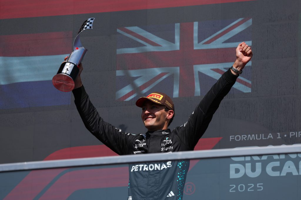 Mercedes driver George Russell celebrates on the podium, raising a trophy with both arms aloft.