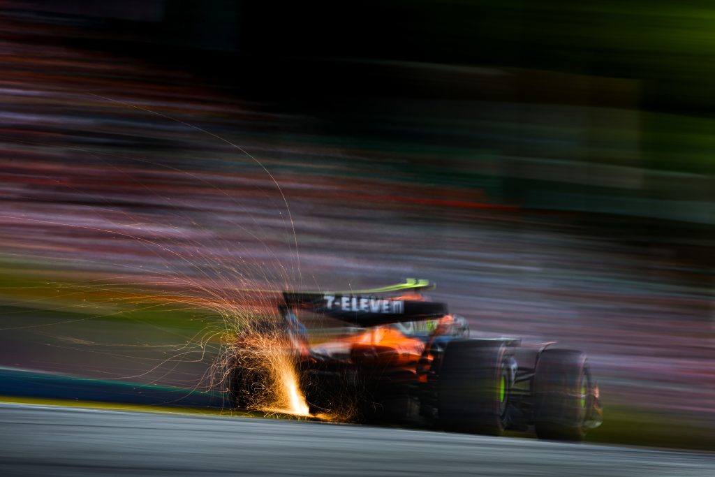Lando Norris’s McLaren (orange car), car #4, charges away from the camera with sparks spraying from the underside as it rides the kerb.