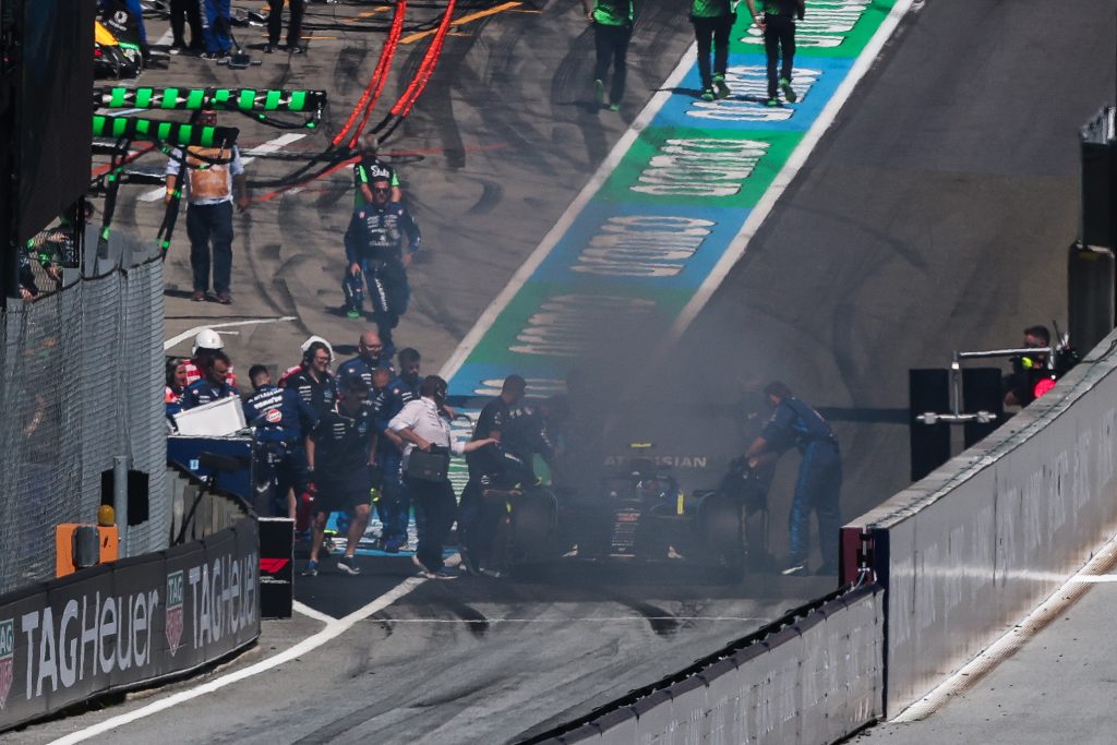 Carlos Sainz’s Williams (dark blue car with white sidepods and blue details), car #55, sits stopped and smoking in the pit lane as crew members and marshals gather around it.