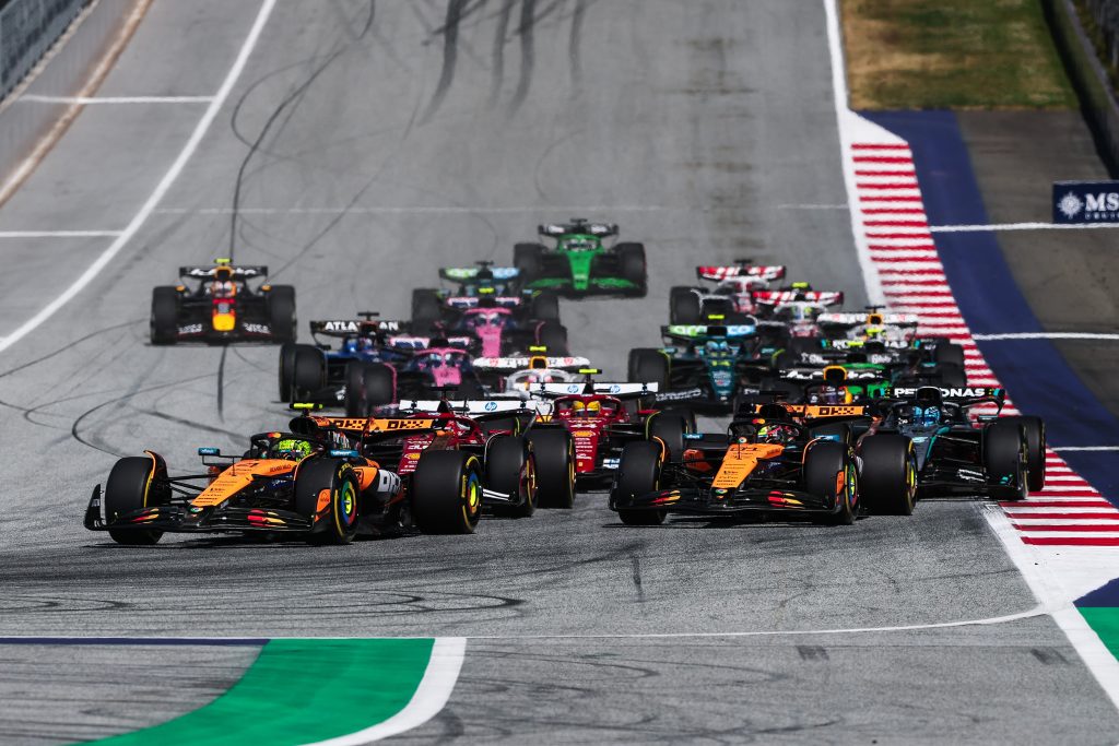 Two McLaren (orange cars) lead the pack into the corner, with Lando Norris’s #4 slightly ahead on the left of Oscar Piastri’s #81, while the lead Ferrari (red car) follows closely behind, the lead Mercedes (silver with black elements and turquoise ...