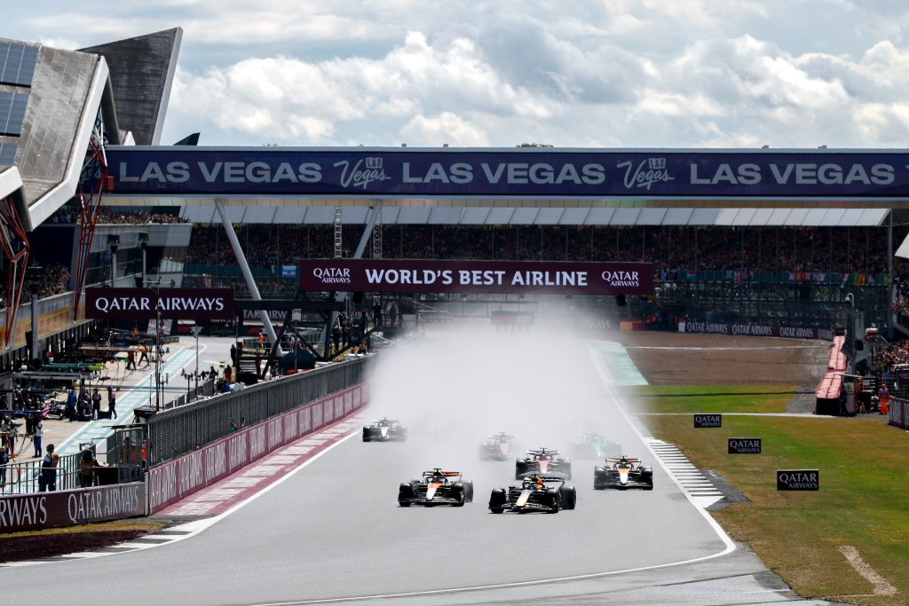 Two McLarens (orange cars), Lando Norris’s #4 and Oscar Piastri’s #81, run side by side through the spray at the start, with Max Verstappen’s Red Bull (dark blue car) just behind and the rest of the field fanning out in their wake.