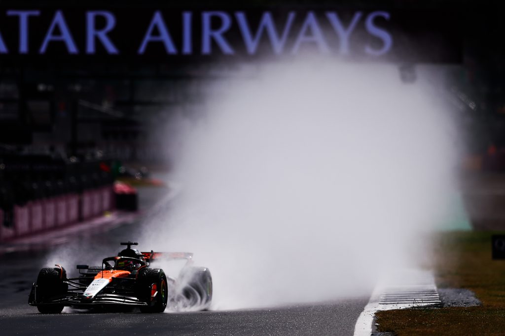 Oscar Piastri’s McLaren (orange car) #81 charges through the wet track at Silverstone, throwing up a huge wall of spray as he runs alone near the edge of the circuit.