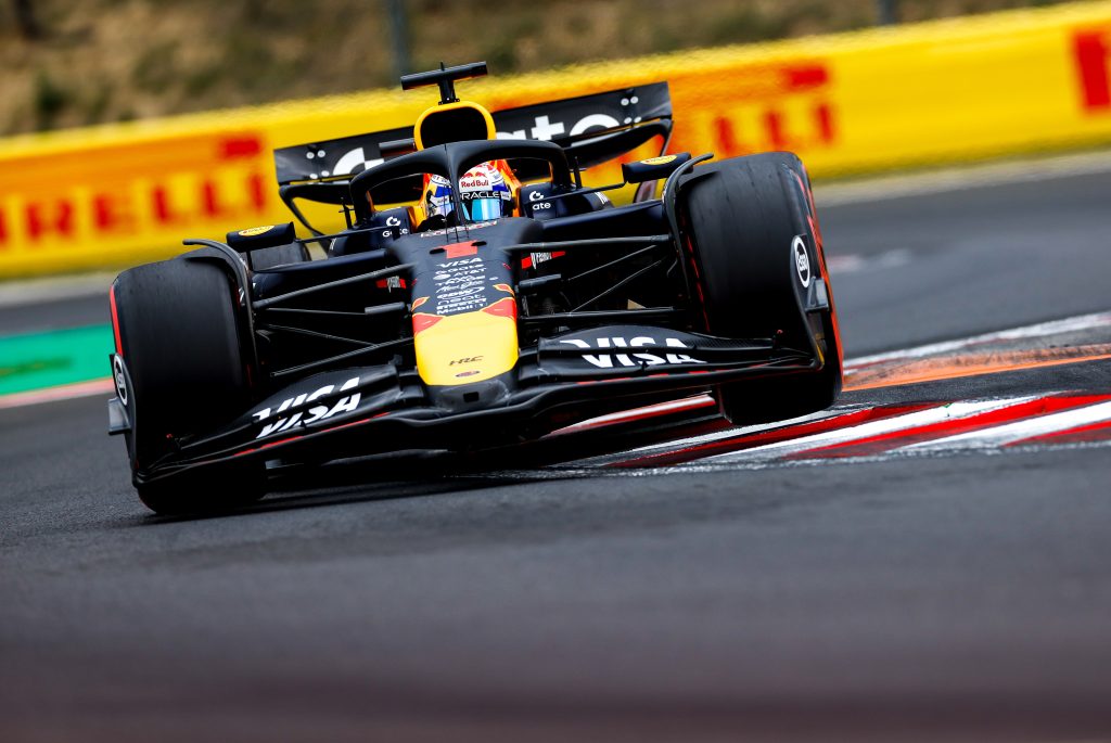 Max Verstappen’s Red Bull #1 (dark blue car with red and yellow details) charges through the corner alone, riding the kerb aggressively as it leans over the apex.