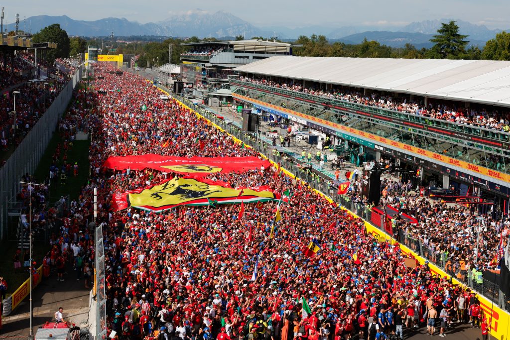 A massive crowd fills the Monza start-finish straight and pit lane during a post-race ceremony, with large Ferrari banners stretched above the fans and grandstands packed on both sides.