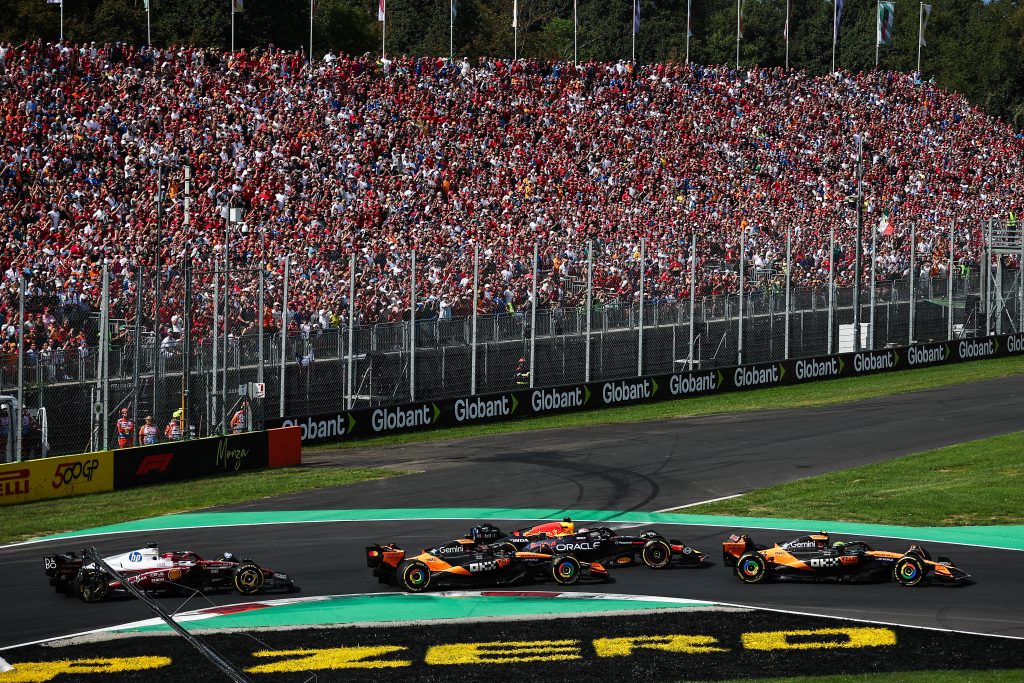 Charles Leclerc’s Ferrari (red car) #16 turns through the inside of the chicane, while Max Verstappen’s Red Bull (dark blue car) #1 runs alongside between the two McLarens: Lando Norris’s McLaren (orange car) #4 to the left and Oscar Piastri’s ...