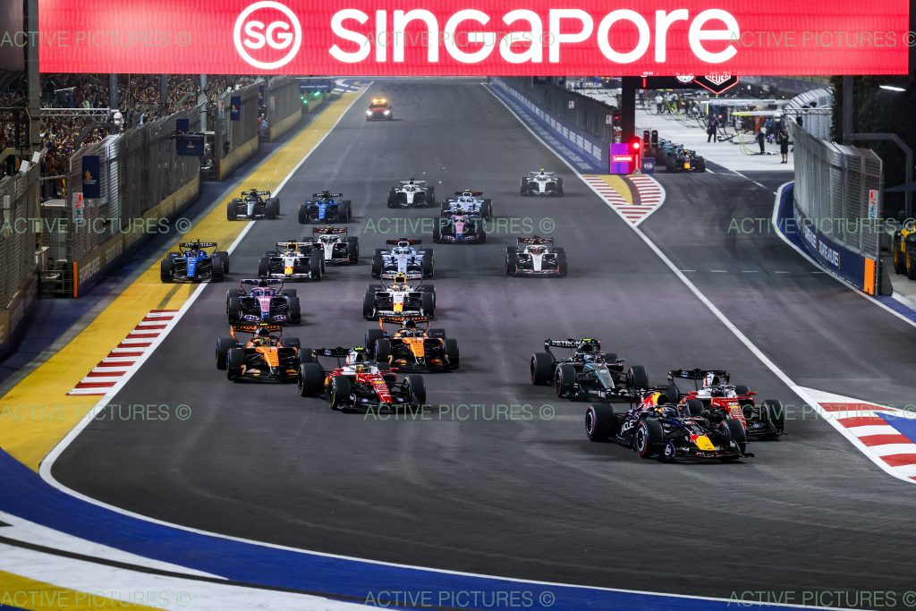 The Red Bull (dark blue car) of Max Verstappen #3 leads the field into Turn 1, while two Ferraris (red cars), Charles Leclerc #16 and Lewis Hamilton #44, fan out behind him, the lead Mercedes (silver and black car) holds the outside line, and two McLar...